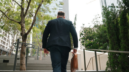 Back view of caucasian manager with formal suit walking up stairs while holding his suitcase in green city. Professional business man going to workiplace while climb stair in eco urban city. Urbane..の写真素材