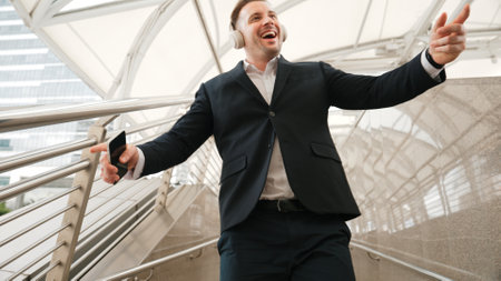Business man dance to relaxing music from headphone at modern corridor. Cheerful project manager wear headset and moving gesture to celebrate successful song with lively mood in urban city. Urbane.の写真素材