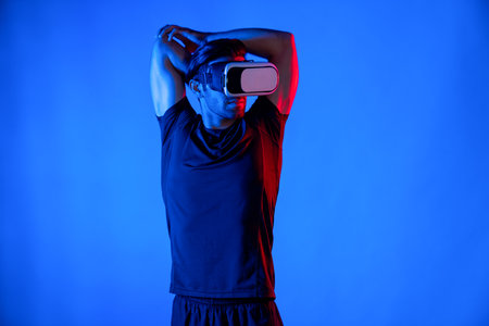 Happy man with VR goggle exercising and stretching arm at neon light background. Caucasian person with casual cloth relaxing and exercising by using virtual reality simulator technology. Deviation.の写真素材
