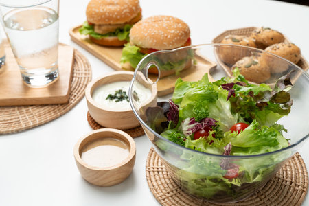 Fresh mix vegetable salad in glass blow beside dish of double meat hamburger and wholegrain bun on meal cooked by homemade serving white table. Preparing special dish menu lists restaurant. Infobahn.の写真素材