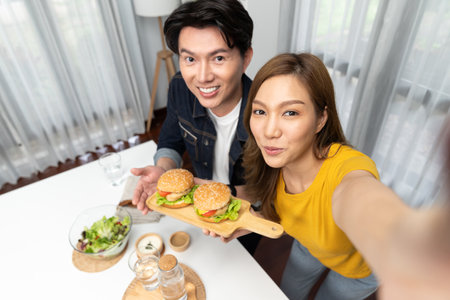 Top view of smiling host chef influencers taking photo with smartphone by selfie holding double hamburger on cooking show special easy with salad side dish homemade on social media channel. Infobahn.の写真素材