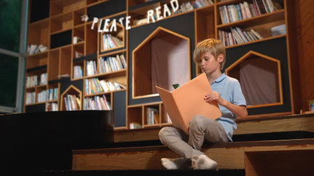 Attractive caucasian clever boy reading a book at library with stack of books placed. Smart child...