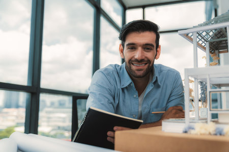 Smiling smart architect engineer looking at camera while sitting near house model. Happy confident businessman wearing casual outfit holding notebook while poses at camera. Civil engineering. Tracery.の写真素材