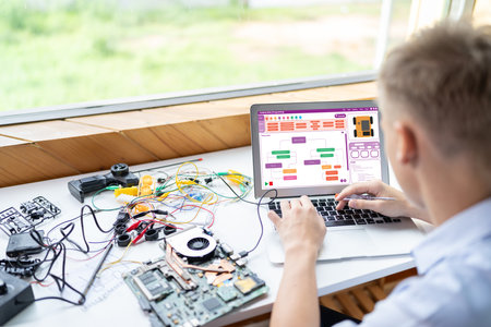 Blonde hair schoolboy in blue shirt code robotics car use laptop learn technology and engineering in STEM class. Around laptop put controller, electric wire, battery charger and laptop. Edification.の写真素材