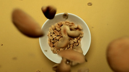 Top view of peanut falling in to white bowl with yellow background. Macrography of a peanut gracefully falling into a white bowl against a vibrant yellow background. Aerial view. Close up. Comestible.の写真素材
