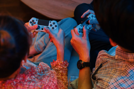 Cropped of hands couple with joysticks sitting sofa playing video game together at back side view...