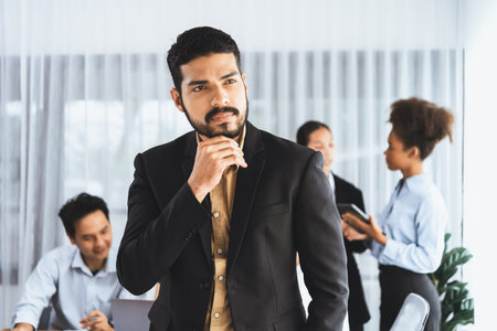 Businessman poses confidently with diverse coworkers in busy meeting room background. Multicultural...
