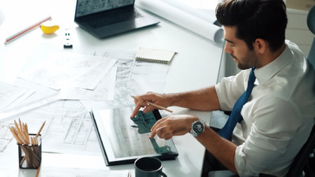 Architect engineer using laptop planning house design at office while sitting at meeting table with blueprint and equipment scatter around.Skilled civil engineer working at blueprint. Alimentation.の写真素材