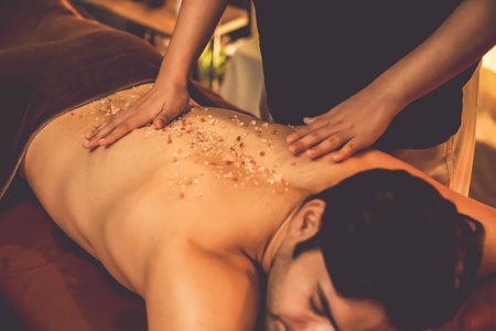 Man customer having exfoliation treatment in luxury spa salon with warmth candle light ambient. Salt scrub beauty treatment in Health spa body scrub. Quiescentの写真素材