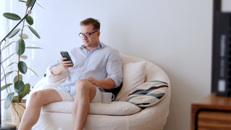Middle age man sit on sofa with coffee cup in room using phone search data for occupation, raising children to be good adult or entertainment. Businessman use technology for convenience. Divergence.の写真素材