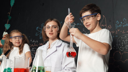 Caucasian boy mixing chemical liquid while teacher giving advice. Professional instructor wearing lab suit looking for diverse student at table with beaker filled with colored solution. Erudition.の写真素材