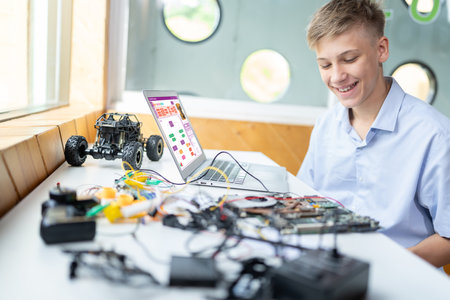 Blonde hair schoolboy in blue shirt watching motherboard while considering and smile in STEM class. On table put laptop, controller, electric wire, battery charger, and robotic vehicle. Edification.の写真素材