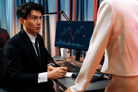 Asian businesswoman standing to discuss with dynamic stock exchange graph with partner to taking...