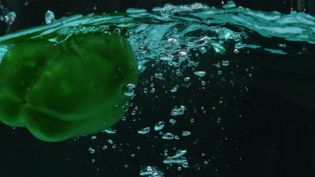Close up of a fresh green bell pepper floating in water. The pepper is round with a green stem at the top. Macro of green bell pepper dropping in to water and splashing with black background. Pabulum.の写真素材