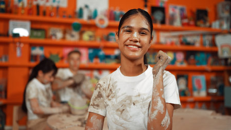 Happy caucasian girl pose at camera while diverse children modeling clay behind. Cute student wearing dirty shirt while looking at camera at workshop in art lesson. Blurring background. Edification.の写真素材