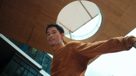 Hispanic dancer looking at camera while dancing at sky scrapper. Attractive handsome hipster moving his hands to hip hop music at modern mall with low angle camera. Outdoor sport 2024. Endeavor.の写真素材