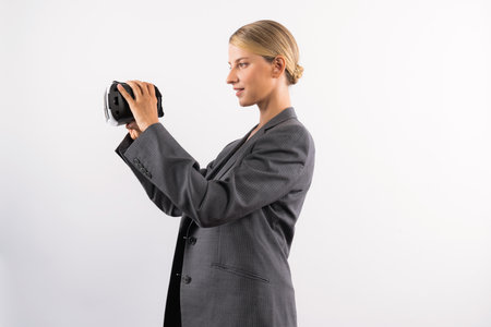 Caucasian business woman holding VR glass while standing at white background. Professional project manager looking visual reality goggle while wearing suit. Innovation technology concept. Contraption.の写真素材