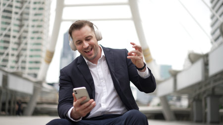 Smart business man wear headphone while sitting at stair in urban city and listen to relaxing music. Happy male leader using headset and moving to song in lively mood surrounded by skyscraper. Urbane.の写真素材