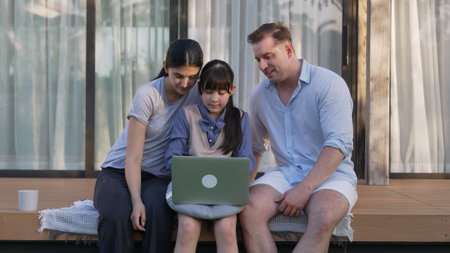 Father, mother and daughter focus laptop at terrace with garden view outside house. Parent use outdoor activity to communicate young generation about environment care cross generation gap. Divergence.の写真素材