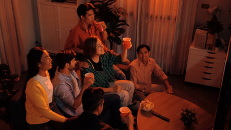 Friend cheering football together and clinking glass at living room. Diverse worker or colleague celebrate winning team while clinking glass and drinking alcohol. Woman clapping hand. Convocation.の写真素材