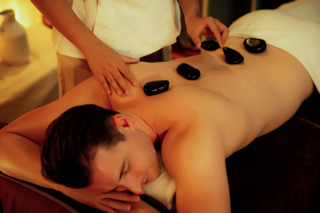 Hot stone massage at spa salon in luxury resort with warm candle light, blissful man customer enjoying spa basalt stone massage glide over body with soothing warmth. Quiescentの写真素材