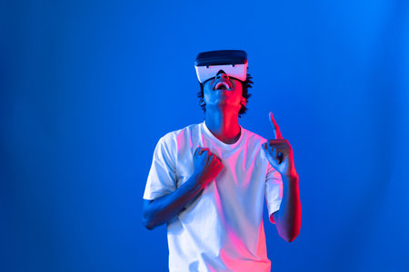 Young African American player wearing VR raising fist up wining game in hologram on blue pink neon lighting wall connecting digital futuristic technology virtual reality metaverse world. Contrivance.の写真素材