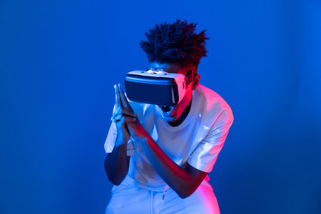 Young African American player using VR turn gun shooting hunter 3D gamer in hologram blue pink neon lighting wall connecting digital futuristic technology virtual reality metaverse world. Contrivance.の写真素材