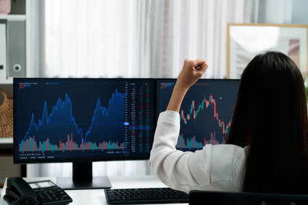Successful young Asian businesswoman earning highest profit with fist up in exchange stock market on dynamic business graph on pc screens at modern office, analyzing financial technology. Stratagem.の写真素材