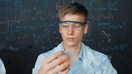 Closeup of boy inspect chemical solution while holding beaker at blackboard with chemical theory. Smart scientist doing experiment while analysis colored solution while wearing lab coat. Edification.の写真素材