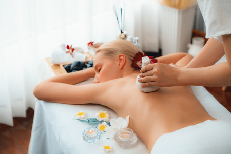 Hot herbal ball spa massage body treatment, masseur gently compresses herb bag on woman body. Tranquil and serenity of aromatherapy recreation in day lighting ambient at spa salon. Quiescentの写真素材