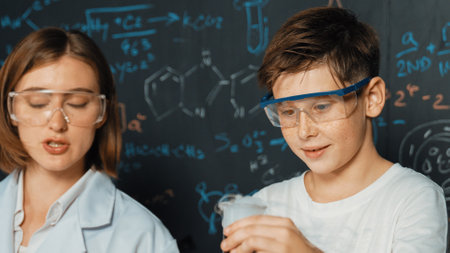 Close up of caucasian boy mixing chemical liquid while teacher giving advice. Instructor wearing lab suit looking for diverse student at table with beaker filled with colored solution. Erudition.の写真素材
