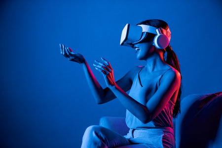 Smart female sitting on sofa surrounded by neon light wear VR headset connect metaverse, futuristic...