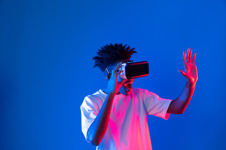 Young African American gaming player wearing VR pointing hologram 3D graphic isolated blue pink neon light wall connecting digital futuristic technology virtual reality metaverse world. Contrivance.の写真素材