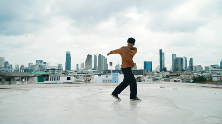 Motion shot of B-boy dance performance by professional street dancer at rooftop with sky scrapper, city view. Attractive asian hipster show energetic footstep. Outdoor sport 2024. Hiphop.の写真素材