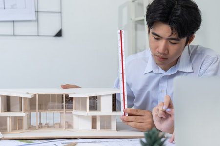 Professional male asian architect using ruler to measure house model length while young beautiful caucasian colleague using laptop to analyzed data on meeting table with house model. Immaculate.の写真素材