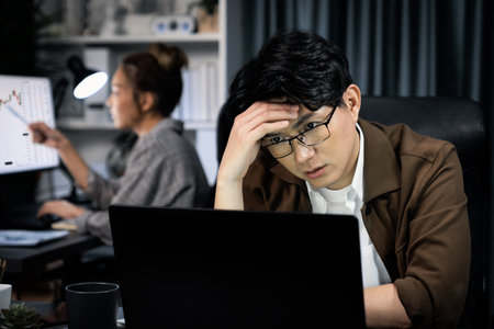 Businessman with stressful face working on laptop waiting email while coworker trading stock...
