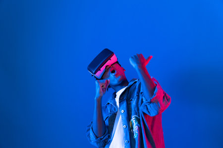 Young African American singer using through VR glasses enjoy singing song with karaoke application on blue neon light wall background in virtual reality metaverse fantasy technology. Contrivance.の写真素材