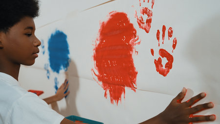 Side view of young children stamp hand with color and paint wall. Highschool student with mixed raced holding tray while paint room with red and blue in art lesson. Creative activity. Edification.の写真素材