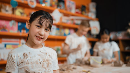 Asian highschool girl look at camera and dirty hand while diverse children modeling clay behind. Happy cute student wearing shirt while looking at camera at workshop. Blurring background. Edification.の写真素材