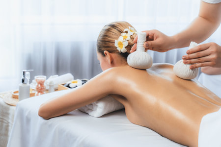 Hot herbal ball spa massage body treatment, masseur gently compresses herb bag on woman body. Tranquil and serenity of aromatherapy recreation in day lighting ambient at spa salon. Quiescentの写真素材