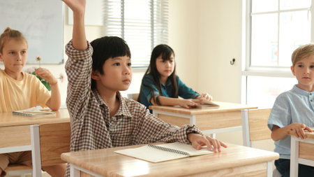 Smart boy raising hand for asking and answering teacher surrounded by multicultural children studying at classroom. Happy student working together, voting, volunteering, calling instructor. Pedagogy.の写真素材