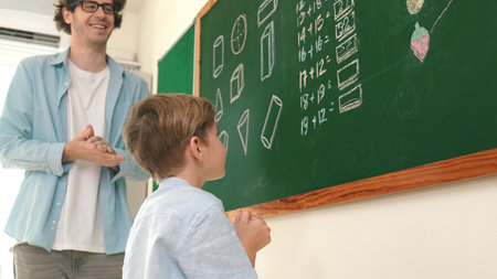 Happy caucasian boy writing answer at blackboard and thinking about theory while standing in front of classroom. Close up of elementary student volunteering to answer question. Education. Pedagogy.の写真素材