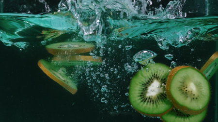 Close up of slices of kiwi fruit falling into a glass of splashed clear cool water. The kiwi slices are green with black seeds and a thin white rind. Some of the slices are curled slightly. Pabulum.の写真素材