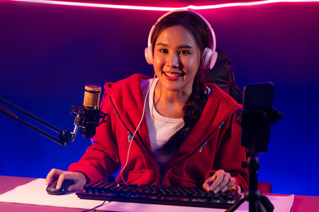 Host channel of smiling beautiful Asian girl streamer playing online game wearing headphone talking with viewers media online recording phone. Esport skilled team players in neon blue room. Stratagem.の写真素材