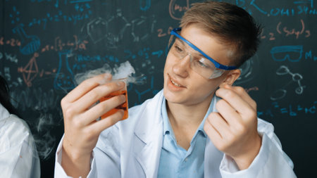 Closeup of boy inspect chemical solution while holding beaker at blackboard with chemical theory....