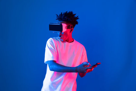 Young African American player wearing VR with using smartphone playing game hologram on blue pink neon light wall connecting digital futuristic technology virtual reality metaverse world. Contrivance.の写真素材