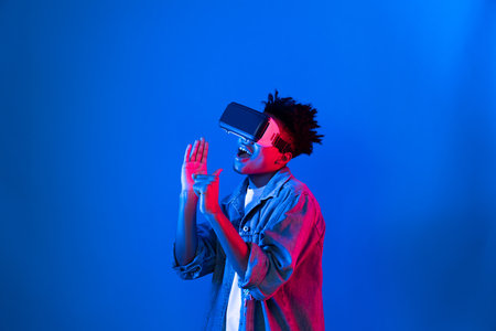 Young African American singer using through VR glasses enjoy singing song with karaoke application on blue neon light wall background in virtual reality metaverse fantasy technology. Contrivance.の写真素材