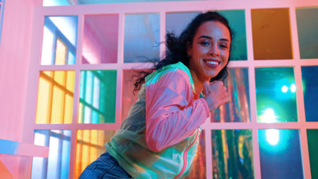 Happy hispanic girl in lively mood moving along music in neon light while wearing colorful cloth at...