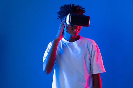 Surprised young African American looking through VR holding interesting object hologram on pink blue neon wall at metaverse world connecting digital futuristic technology virtual reality. Contrivance.の写真素材
