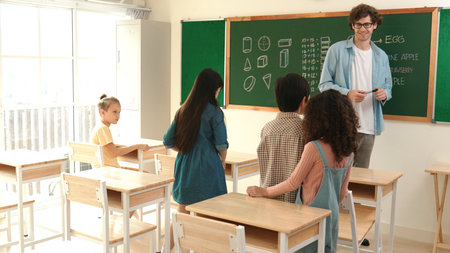 Diverse elementary student walking in classroom while teacher looking at child. Cute children attend...